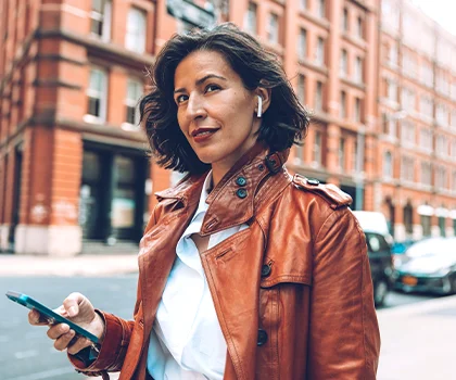 Brunette woman in orange leather coat, walking through the city with headphones and cellphone - Brow Lift in San Francisco, CA