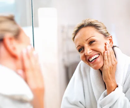 Smiling blonde woman in rob, hand resting on her face, looking in the mirror - Neck and Chin Liposuction in San Francisco, CA