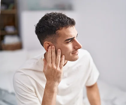 Profile of a brunette man in white shirt, sitting on bed with hand touching ear - Otoplasty in San Francisco, CA