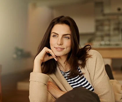 brunette woman in her living room, wearing striped shirt and beige cardigan, smiling with chin resting on hand - PRP Hair Treatment in San Francisco - CA