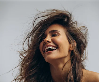 Brunette woman against a grey background, smiling with hair in the wind - Rhinoplasty in San Francisco, CA